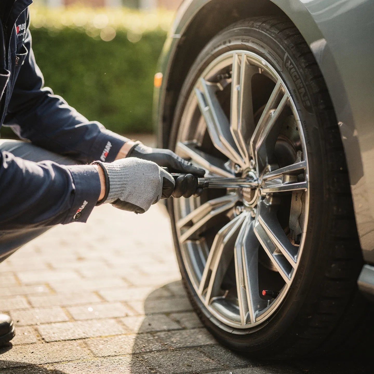 Technicien pneumatique en combinaison de travail et gants de protection utilisant une clé en croix pour desserrer les boulons d'une roue sur véhicule premium, pavés suisses au sol